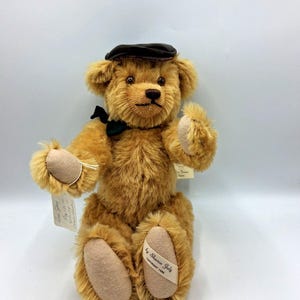 May include: A golden brown teddy bear wearing a dark brown cap and a black bow tie. The bear has tan paw pads and a tag that reads "by Sharon July Copyright 1986".