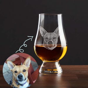 May include: A clear glass whisky tumbler with a dog's face etched into the side. The dog is a brown and white terrier mix. The glass is filled with amber liquid.