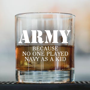May include: A clear, square-shaped rocks glass filled with amber liquid. The glass features white text that reads "ARMY" above the phrase "BECAUSE NO ONE PLAYED NAVY AS A KID."