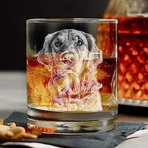 May include: A clear rocks glass filled with amber liquid, featuring an etched portrait of a dog. The dog's image is detailed, with visible fur and facial features. The glass sits on a dark gray napkin, with a blurred background.