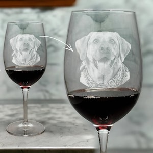 May include: Two clear wine glasses filled with red wine. Each glass has an etched image of a dog's face. The dog wears a bandana. The wine glasses are on a marble surface.