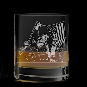 May include: A clear glass tumbler with a design etched into the side. The design features four people, three of whom are holding an American flag. The tumbler is filled with a brown liquid.