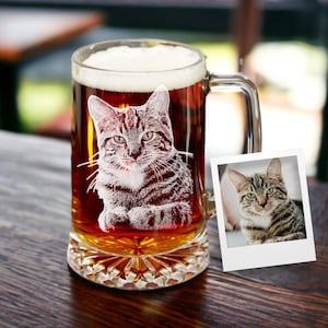 May include: A clear glass beer mug filled with beer and foam, featuring an etched image of a tabby cat. A small photo of the cat is next to the mug. The mug sits on a wooden surface.