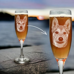 May include: Two champagne flutes filled with a light brown liquid, each etched with a detailed dog portrait. The flutes have a clear bowl, a white stem with an orange accent, and a silver base. The background shows a blurred sunset over water.
