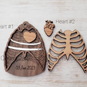 May include: Two wooden ornaments shaped like a rib cage and a heart. The heart ornament has a smaller heart cutout in the center. The rib cage ornament has a cutout design. The heart ornament has the text "03 Jan 2023" inscribed on it. The ornaments are labeled "Heart #1" and "Heart #2".