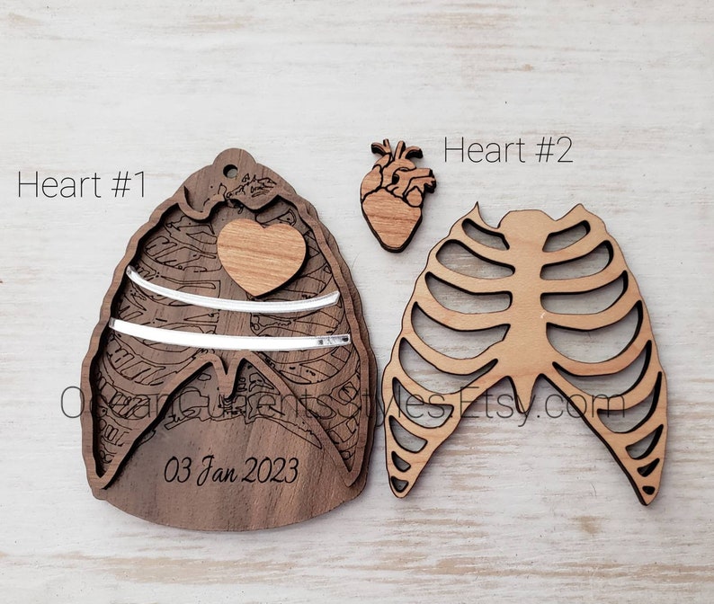 May include: Two wooden ornaments shaped like a rib cage and a heart. The heart ornament has a smaller heart cutout in the center. The rib cage ornament has a cutout design. The heart ornament has the text "03 Jan 2023" inscribed on it. The ornaments are labeled "Heart #1" and "Heart #2".