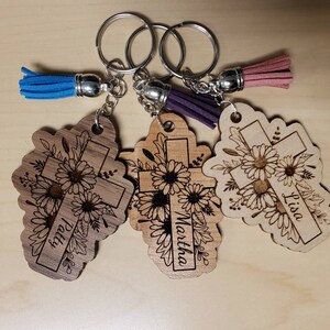 May include: Three wooden cross keychains with floral designs and personalized names. Each keychain has a different colored tassel: blue, purple, and pink. The crosses are engraved with the names Patty, Martha, and Lisa.