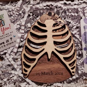 May include: A wooden ribcage ornament with a detailed design and the date "25 March 2024" engraved on the bottom. The ornament is surrounded by white packing peanuts.