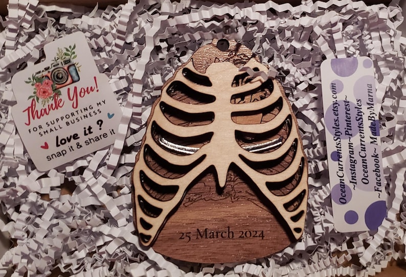 May include: A wooden ribcage ornament with a detailed design and the date "25 March 2024" engraved on the bottom. The ornament is surrounded by white packing peanuts.