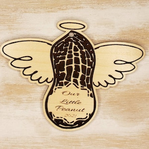 May include: Wooden angel ornament shaped like a peanut with a halo and wings. The peanut has a textured surface and the text "Our Little Peanut 2021!" is engraved on the bottom.