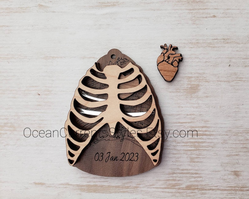 May include: Wooden ornament shaped like a ribcage with a small heart ornament beside it. The ribcage ornament has the date "03 Jan 2023" engraved on it.