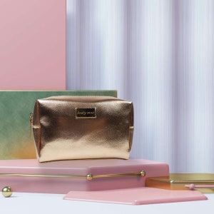 May include: A gold metallic makeup bag with the text "dusty rose" on the front. The bag is sitting on a pink platform.