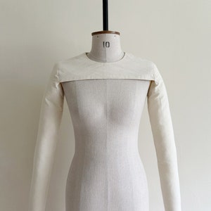 May include: A white fabric garment with long sleeves and a round neckline. The garment is displayed on a white dress form.
