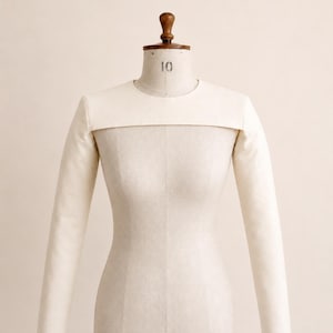 May include: A cream-colored bolero shrug featuring long, fitted sleeves and a high, rounded neckline. The garment is displayed on a mannequin torso, showcasing a structured, minimalist design that covers the shoulders and upper chest area.