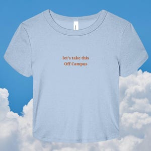 May include: A light blue short-sleeved t-shirt with a crew neckline. The center of the chest features the text "let's take this Off Campus" printed in a dark orange font. The garment has a standard fit with a straight hemline at the bottom.