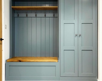 May include: A gray painted wooden entryway bench with a built-in storage compartment and a shelf above. The bench has a natural wood top and a set of hooks for hanging coats or bags. The bench is attached to a tall cabinet with two doors.