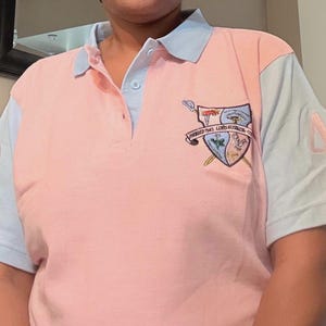 May include: A short-sleeve polo shirt featuring a color-blocked design with a pink center panel and light blue sleeves and collar. The shirt displays a crest on the left chest and the number 49 in pink on the right sleeve, with a button-up placket.