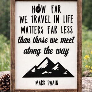 May include: A wooden framed sign featuring the quote "How far we travel in life matters far less than those we meet along the way" and a mountain graphic. The quote is attributed to Mark Twain. The sign is displayed outdoors.