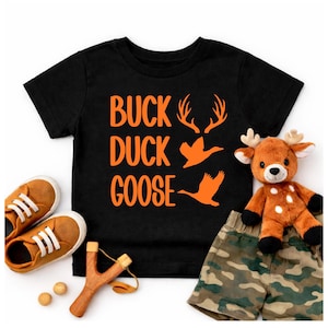 May include: Black t-shirt with orange text and graphics that read "BUCK DUCK GOOSE". The shirt is paired with brown sneakers, a wooden slingshot, wooden balls, camouflage shorts, and a stuffed deer.
