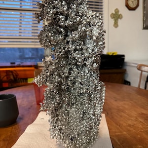 May include: A silver-colored, textured sculpture with a complex, organic form. The sculpture is made of many small, interconnected pieces, creating a porous, almost cellular appearance. It sits on a white paper towel on a wooden table.