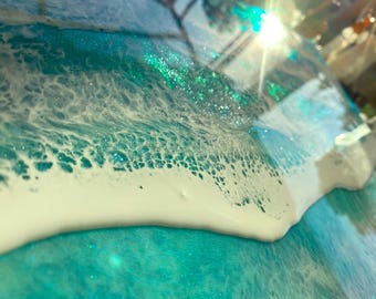 May include: Close-up of a teal blue and white resin art piece. The piece has a wave-like design with white foam and glitter embedded in the resin.