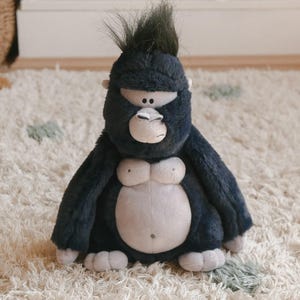 May include: A plush gorilla toy featuring dark charcoal-coloured fur, a light grey face, chest, and belly, and a tuft of dark hair on top of its head. The stuffed animal has small black eyes, a rounded nose, and soft, light grey hands and feet.