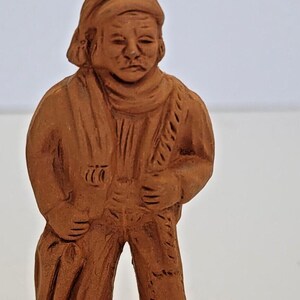 May include: A terracotta figurine of a person wearing a hat and traditional clothing. The figurine is standing with its hands clasped in front of it.