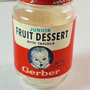 Peut inclure: Un pot de nourriture pour bébé Gerber vintage. Le pot a une étiquette rouge et blanche avec les mots "Junior Fruit Dessert with Tapioca" et un visage de bébé. Le pot est étiqueté "Poids net 220g."