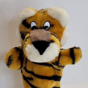 May include: A plush tiger hand puppet with orange and black stripes. The puppet has white fur around the eyes and nose, and black eyes with white pupils.
