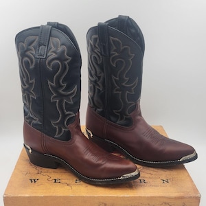 May include: A pair of two-tone cowboy boots featuring dark brown leather vamps and black leather shafts with decorative stitching. The boots include silver-tone metal toe and heel guards and are displayed on a Laredo Western Boots cardboard box.