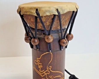 May include: A small, brown wooden drum with a leather head and black rope lacing. The drum has a carved design on the side and is decorated with brown wooden beads.