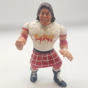 Può includere: Un action figure in plastica di un wrestler con capelli castani, che indossa una maglietta bianca con la scritta rossa Hot Rod, un kilt a quadri rossi e bianchi, fasce rosse per le braccia e stivali da wrestling neri con lacci bianchi.