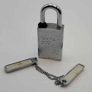 May include: A silver-colored padlock with a curved shackle and the text "IBERET 500" on the front. Two matching keys are connected by a chain. The keys have a rectangular shape with a light-colored insert.