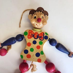 May include: Wooden pull-string clown toy with a painted face, brown hair, and a red nose. The clown's body is a light wood color with a red bow tie and colorful polka dots. Arms and legs are blue and red.