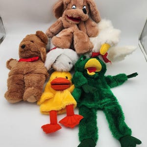 May include: A collection of plush animal puppets, including a brown dog, a white rabbit, a yellow duck, a green bird, and a larger brown dog puppet. The puppets have various colors and textures, with red accents on some.