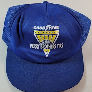 May include: A blue trucker hat with a mesh back and a curved brim. The front of the hat features the Goodyear logo and the text "PERRY BROTHERS TIRE 60 YEARS" in white and yellow.