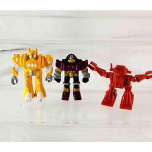 May include: Three small robot figurines in yellow, purple, and red. The yellow robot has white and silver accents. The purple robot has black and yellow accents. The red robot has articulated arms and legs. All three are standing upright.