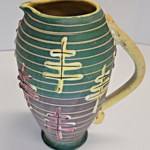 May include: A green and yellow ceramic pitcher with a stylized design of branches and leaves. The pitcher has a wide mouth and a handle.