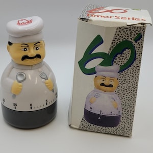 Könnte beinhalten: Ein neuartiger Küchenwecker in Form eines Kochs, der einen weißen Hut und eine Uniform trägt und einen Löffel und eine Gabel hält. Der Timer hat einen schwarzen Sockel mit einem nummerierten Zifferblatt. Die daneben stehende Box hat ein ähnliches Kochbild und den Text "60 TimerSeries".