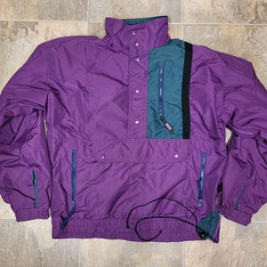 May include: A purple anorak-style jacket with a high collar and multiple pockets. The jacket features a teal and black accent panel on the chest, with blue zippers and a drawstring hem. The jacket is made of a lightweight, water-resistant material.
