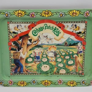 Op de afbeelding: Een vintage groene metalen dienblad met een kleurrijke illustratie van Cabbage Patch Kids. De dienblad heeft een bloemenrand en het Cabbage Patch Kids-logo. De illustratie toont kinderen in een veld met een boom en een rode banner.