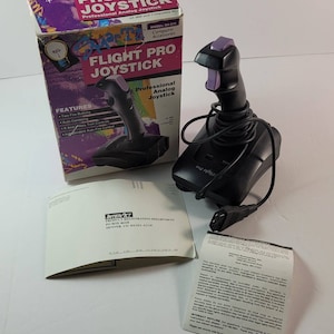 Puede incluir: Un joystick Flight Pro negro con detalles morados, junto a su caja original. La caja muestra el nombre del producto y sus características. Incluye documentos y un cable conector.