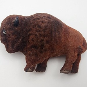May include: A brown, plush bison figurine with a textured surface. The bison has small black horns and a single visible eye. The figurine is standing on four legs and is a detailed representation of the animal.
