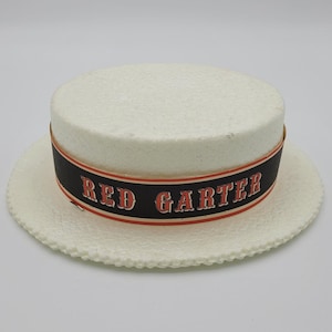 May include: A white straw boater hat with a black band displaying the text "RED GARTER" in red. The hat has a round crown and a wide brim with a scalloped edge. A classic hat, often worn in summer.