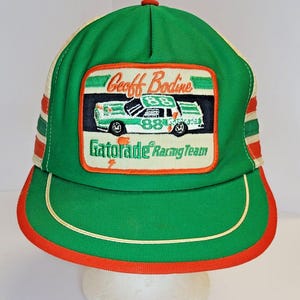 May include: Green and white trucker hat with a red and white stripe on the side. The hat has a patch on the front that says "Geoff Bodine Gatorade Racing Team" and features a green and white race car with the number 88 on it.