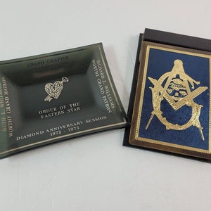 May include: Two Masonic items: a dark green ashtray with text and a heart-shaped emblem, and a blue and gold emblem on a black background. The ashtray reads "Order of the Eastern Star" and "Diamond Anniversary Session 1972-1973".