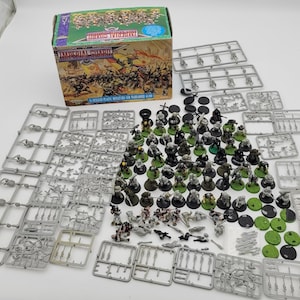 May include: A box set of Imperial Guard miniatures for Warhammer 40,000, with various plastic sprues, bases, and assembled figures. The box features artwork of a battle scene. The miniatures are in various colors, including green, gray, and black.