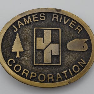 May include: Oval-shaped, bronze-colored metal belt buckle with the words "JAMES RIVER CORPORATION" in a circular pattern. The center features a stylized tree, a geometric design, and a duck-like shape.