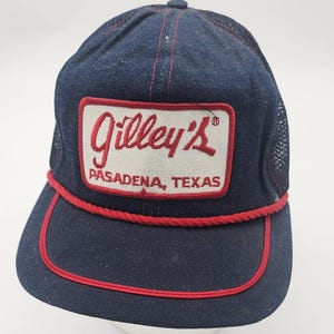 May include: A dark blue denim trucker hat featuring a rectangular white patch with red embroidered text that reads Gilley's PASADENA, TEXAS. The cap includes a red braided cord across the base of the crown and a matching red trim on the brim.