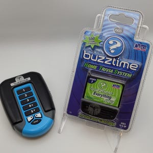 Könnte beinhalten: Ein Buzztime Home Trivia System Spielset. Das Set enthält einen schwarz-blauen Handheld-Spielcontroller und eine Trivia-Spielkassette in der Originalverpackung. Die Verpackung trägt die Aufschrift "buzztime" und "Home Trivia System".